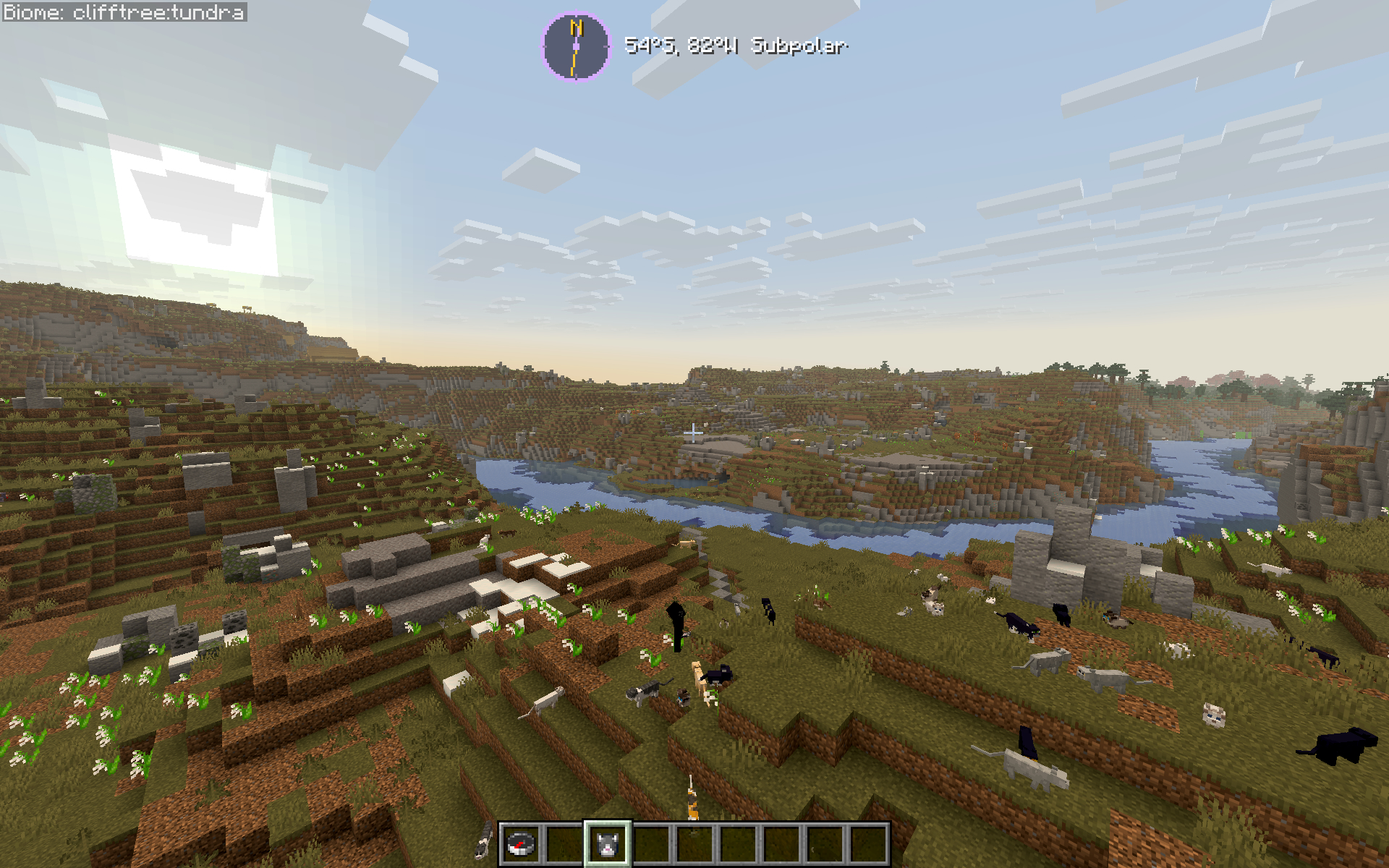 A Minecraft screenshot showcasing a sprawling landscape generated by the Latitude mod. A wide, blue river winds through a rolling green and dirt valley, stretching toward steep cliffs with scattered trees under a bright, clear sky. In the immediate foreground, a grassy hillside is unexpectedly populated by over a dozen cats of various breeds (black, tuxedo, and tabby). At the top center of the screen, the mod's custom HUD element displays the location readout: "54°S, 82% Subpolar". The player's hotbar is visible at the bottom of the frame, showing a compass and an item slot.