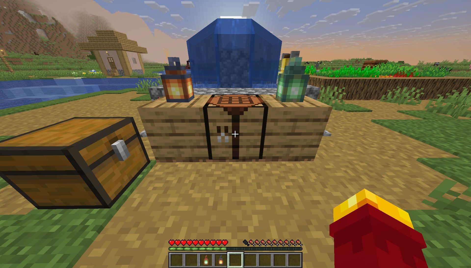 crafting table in front of the plain village fountain, an oak block with a lantern on either side, copper and iron, chest on the left, a hill behind the village on the left, almost sunset, you can see the sun setting behind the fountain
