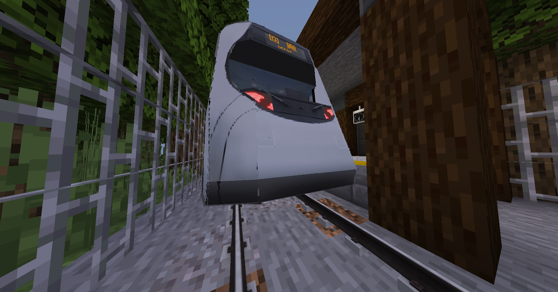 The train has a sleek, futuristic design with visible headlights and a digital destination display on the front. The surrounding environment includes rails, iron fencing, and vegetation, giving the scene the appearance of a train station set within a forest or rural area.